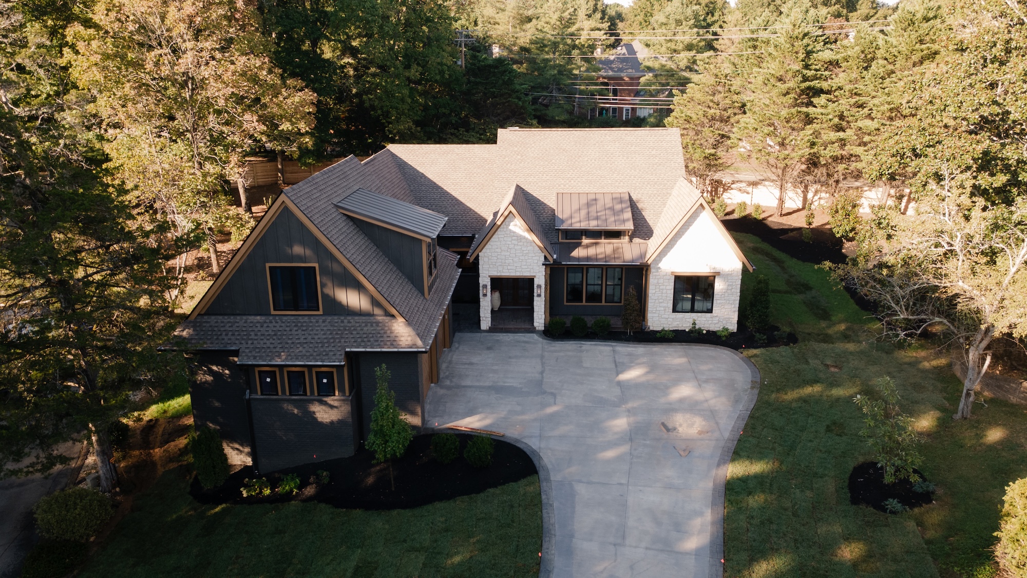 Aerial view of a modern house with a large concrete driveway, landscaped front yard, and surrounded by trees.