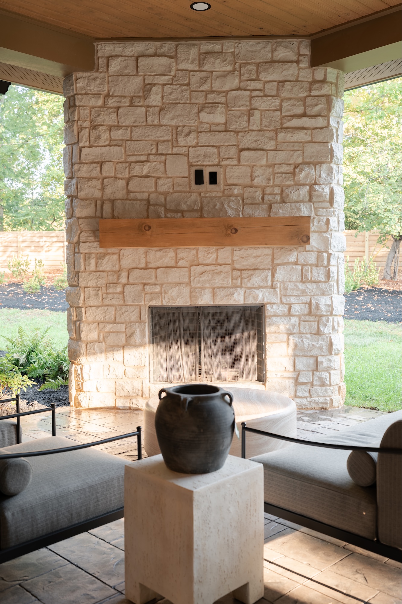 Outdoor covered patio with stone fireplace featuring a wooden mantel, surrounded by two cushioned gray lounge chairs and a round table, with a black pottery vase on a square stone pedestal in the foreground.