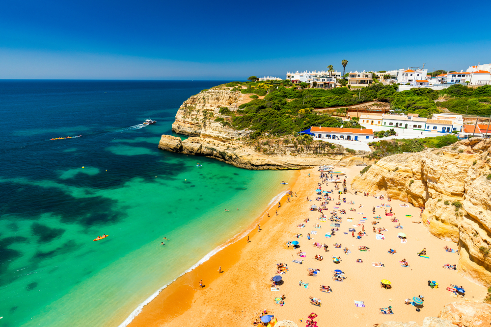 A crowded beach in the Algarve, with turquoise water and cliffs under a clear blue sky