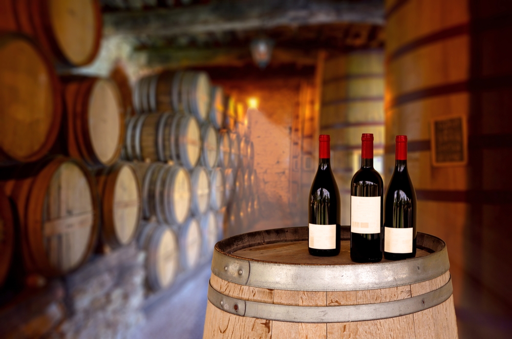 Three red wine bottles on a wooden barrel in a cellar filled with aging wine casks