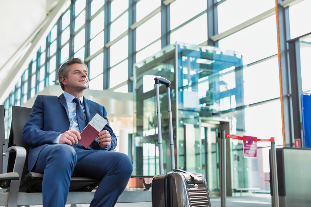 Businessman waiting in an airport lounge holding his passport and boarding pass with luggage nearby