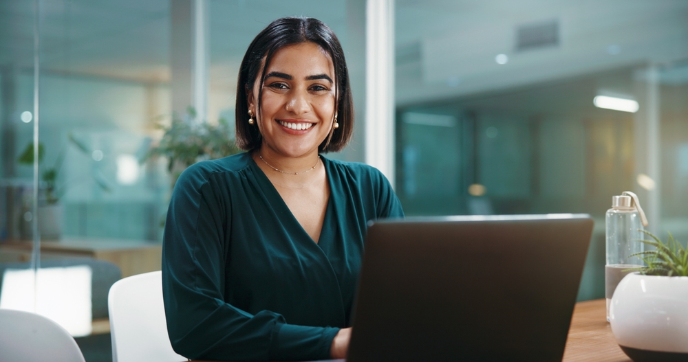 A professional woman smiling while working on a laptop in a bright, modern office