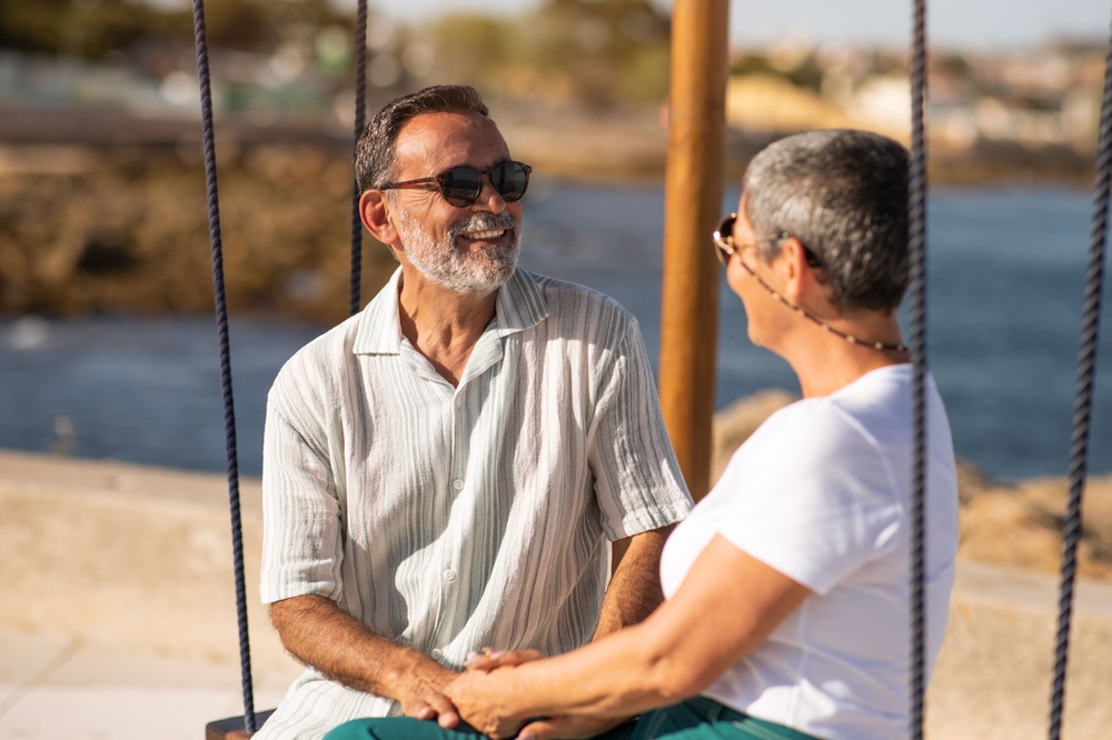 A happy mature couple sits on a seaside swing set, smiling and holding hands