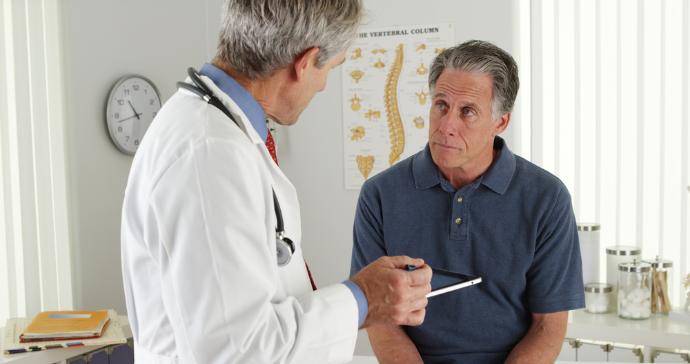 Male doctor in a white lab coat using a tablet to consult with a senior patient in a clinic