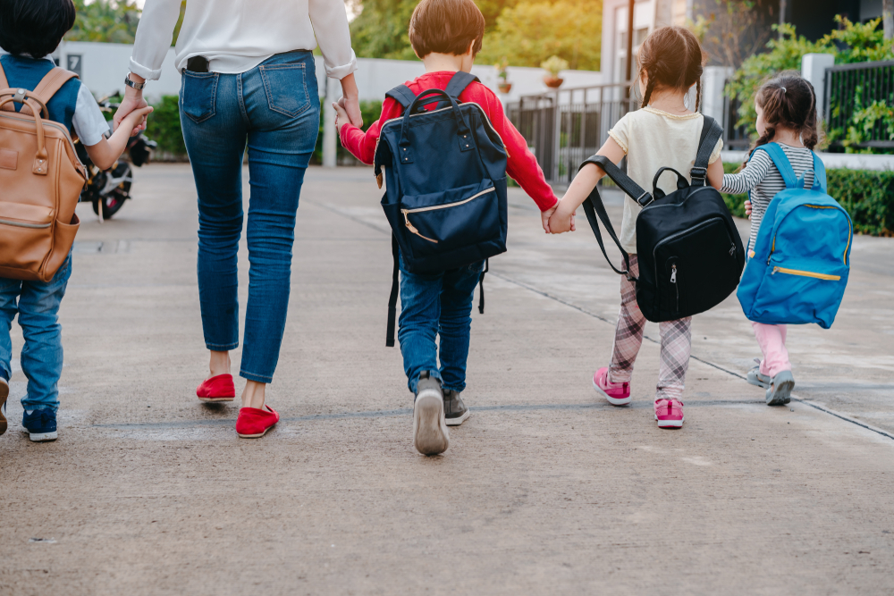 Four children with backpacks walking and holding hands with an adult on a paved path