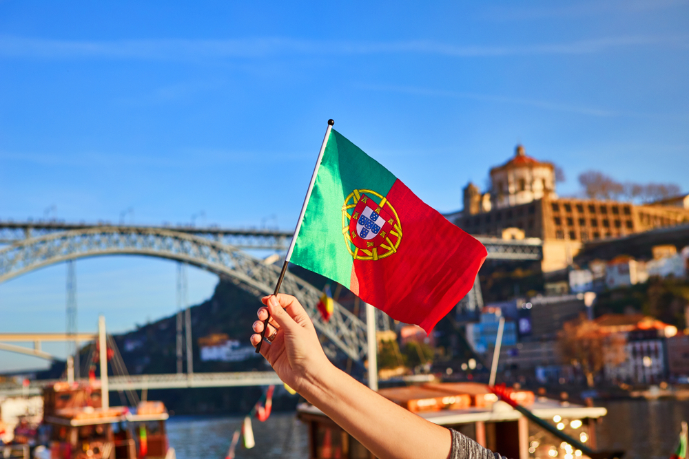 A hand waves a small Portuguese national flag in front of Dom Luís I Bridge in Porto