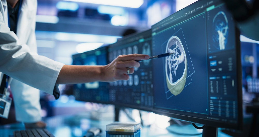 A medical professional points a pen at a brain scan displayed on a large computer monitor