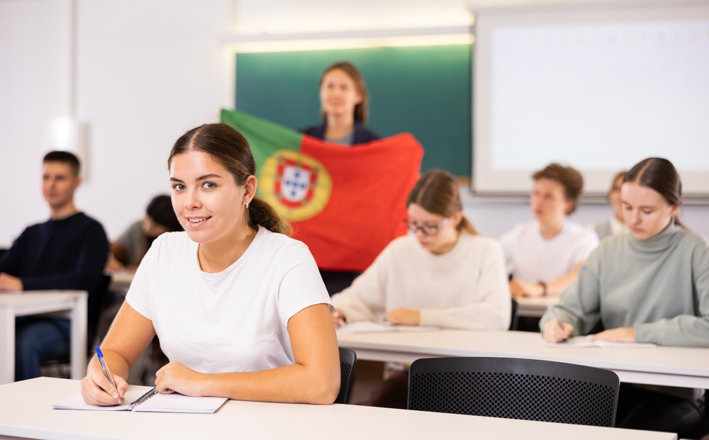 A young woman, with a Portuguese flag behind her, is writing in a classroom