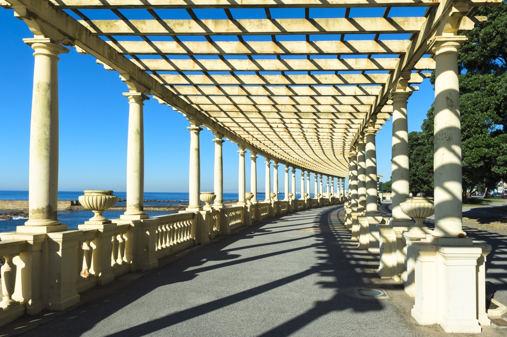 A long white stone pergola walkway next to the ocean under a clear blue sky