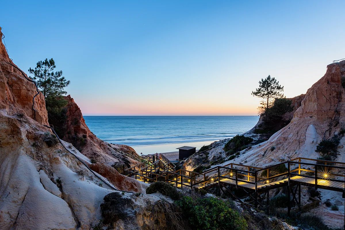 Illuminated wooden boardwalk winding down red cliffs to Praia da Falésia beach at sunset