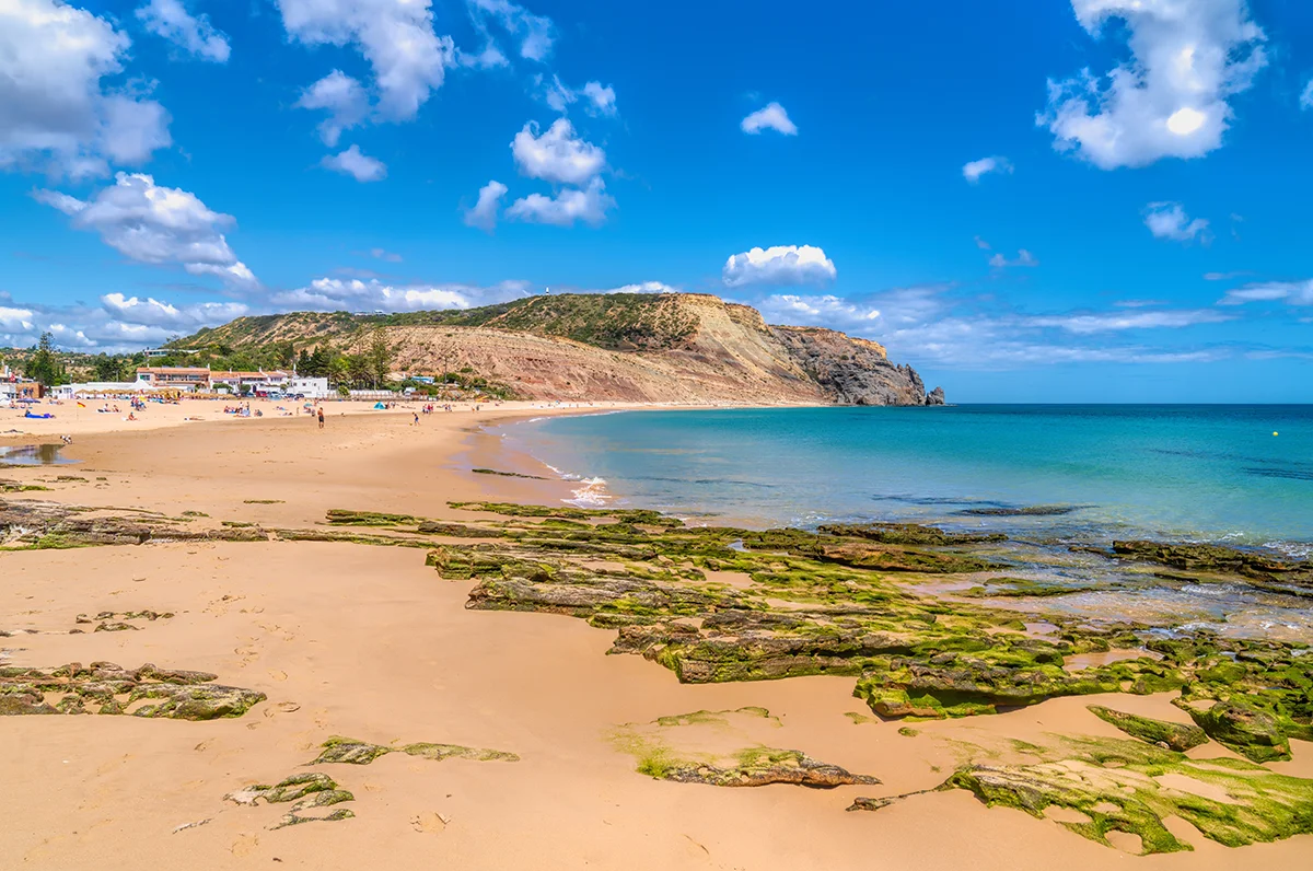 Mossy rocks line a wide, golden sand beach under a bright blue sky with fluffy white clouds