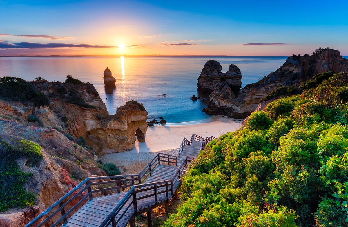 A wooden staircase leads down to a serene sandy cove as the sun rises over the ocean