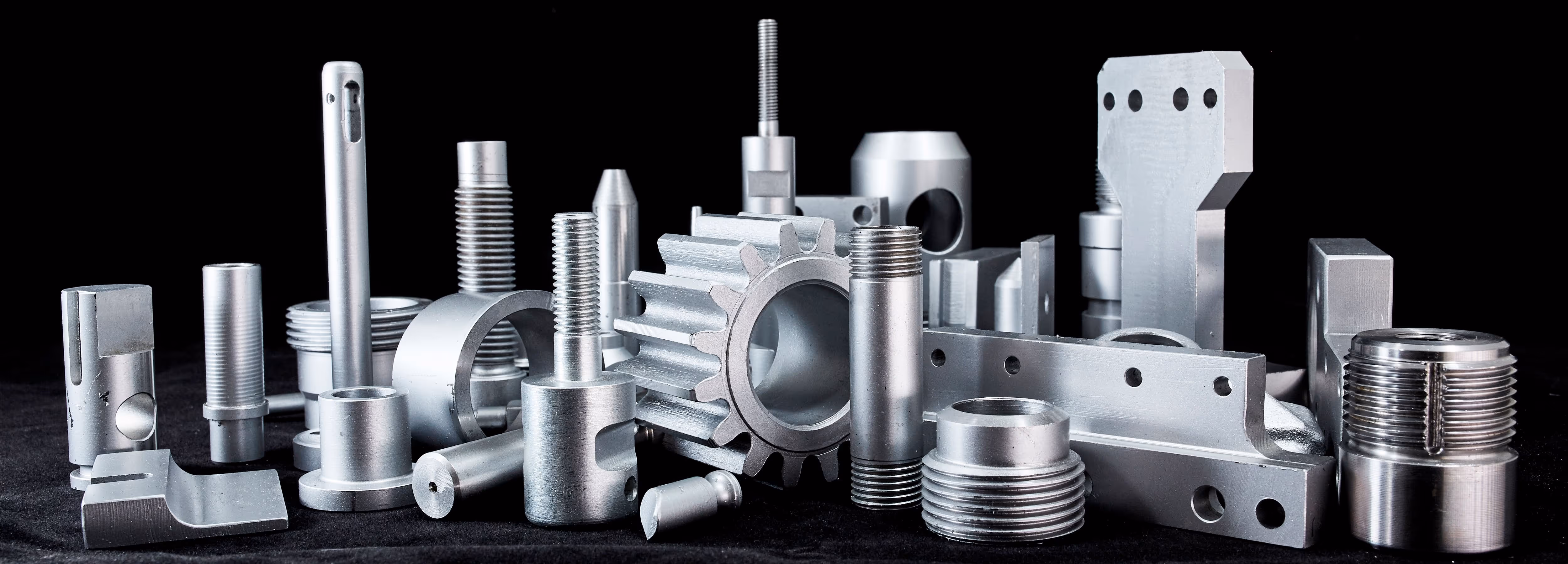 Assorted precision CNC machined metal parts including gears, threaded components, and cylindrical shapes on a black surface.
