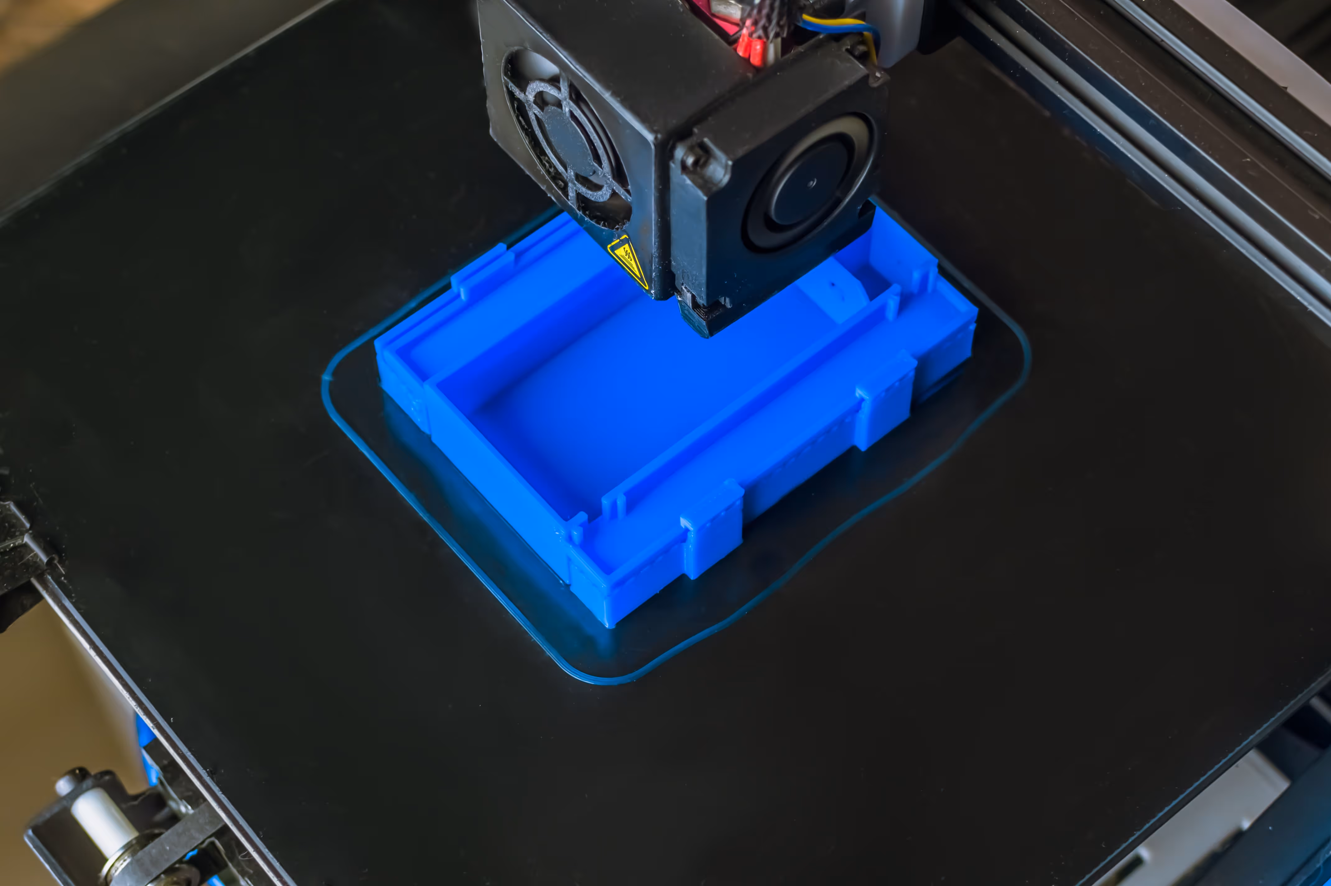 3D printer creating a blue rectangular plastic prototype on a black printing bed.