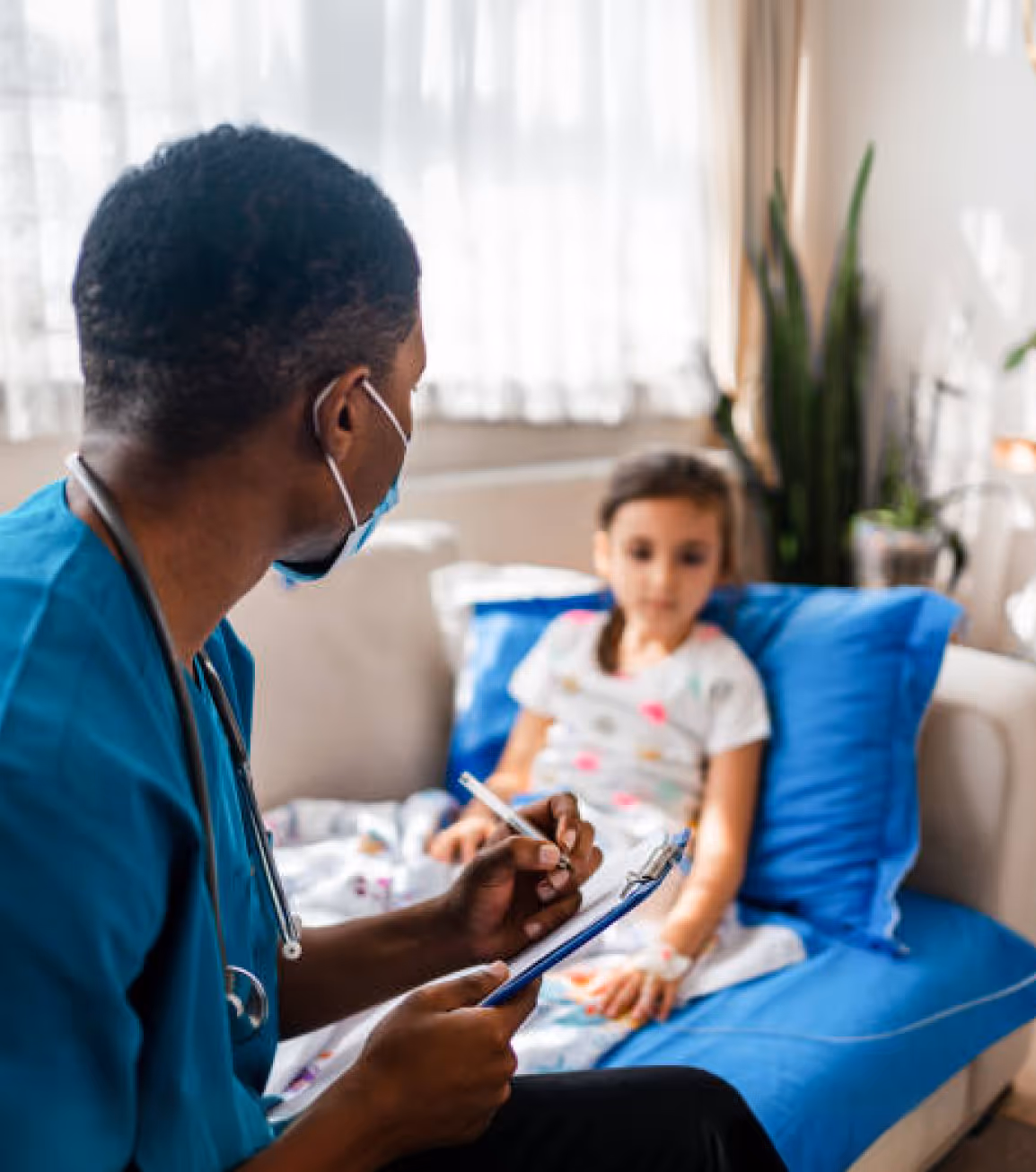 Healthcare professional wearing mask and scrubs talking to a young girl sitting on a couch with an IV in her hand.