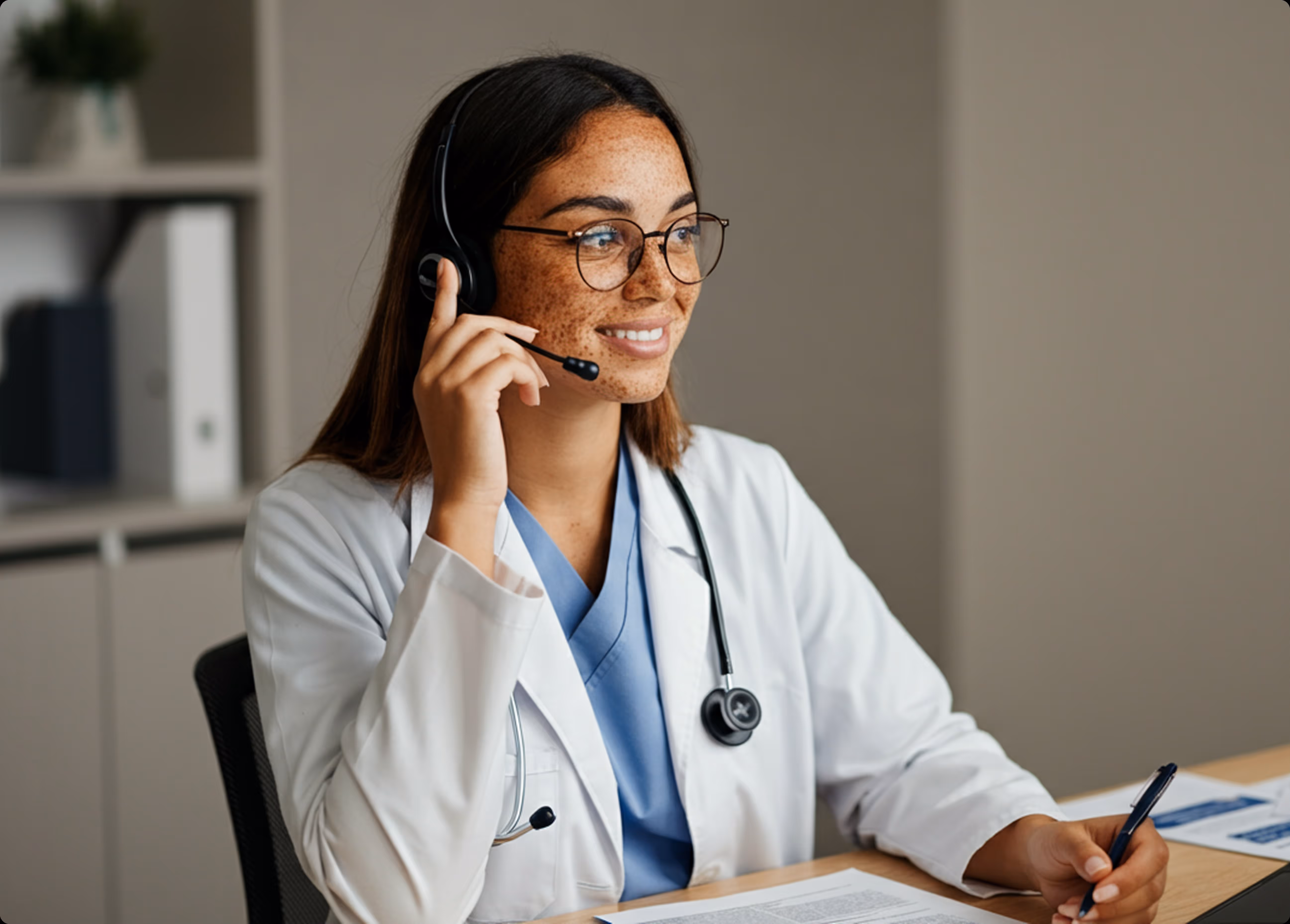 Smiling female doctor wearing a headset and glasses, sitting at a desk with medical documents, holding a pen.