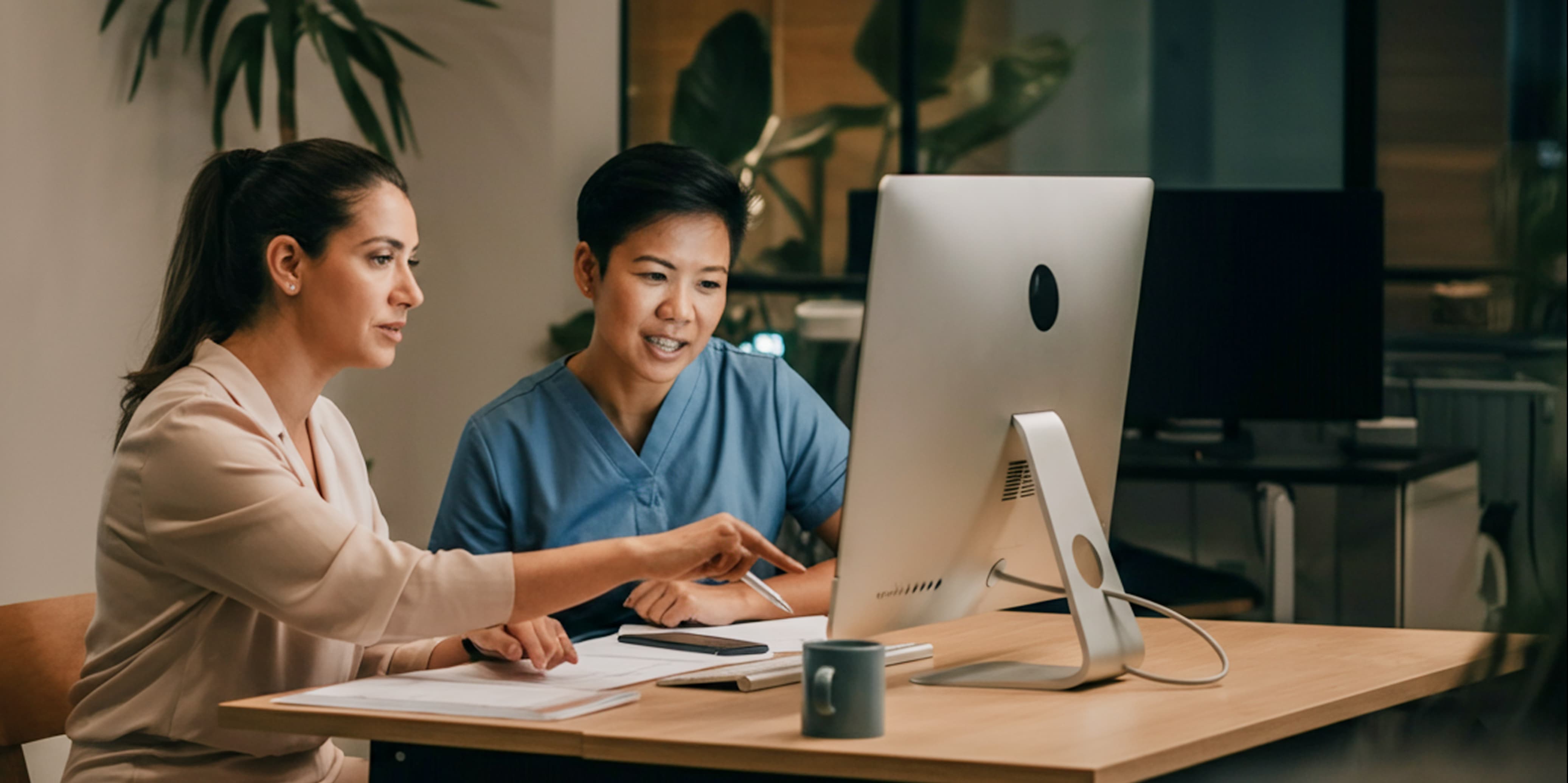 Two women working together at a desk, looking at a computer screen; one woman points at the screen while the other listens.