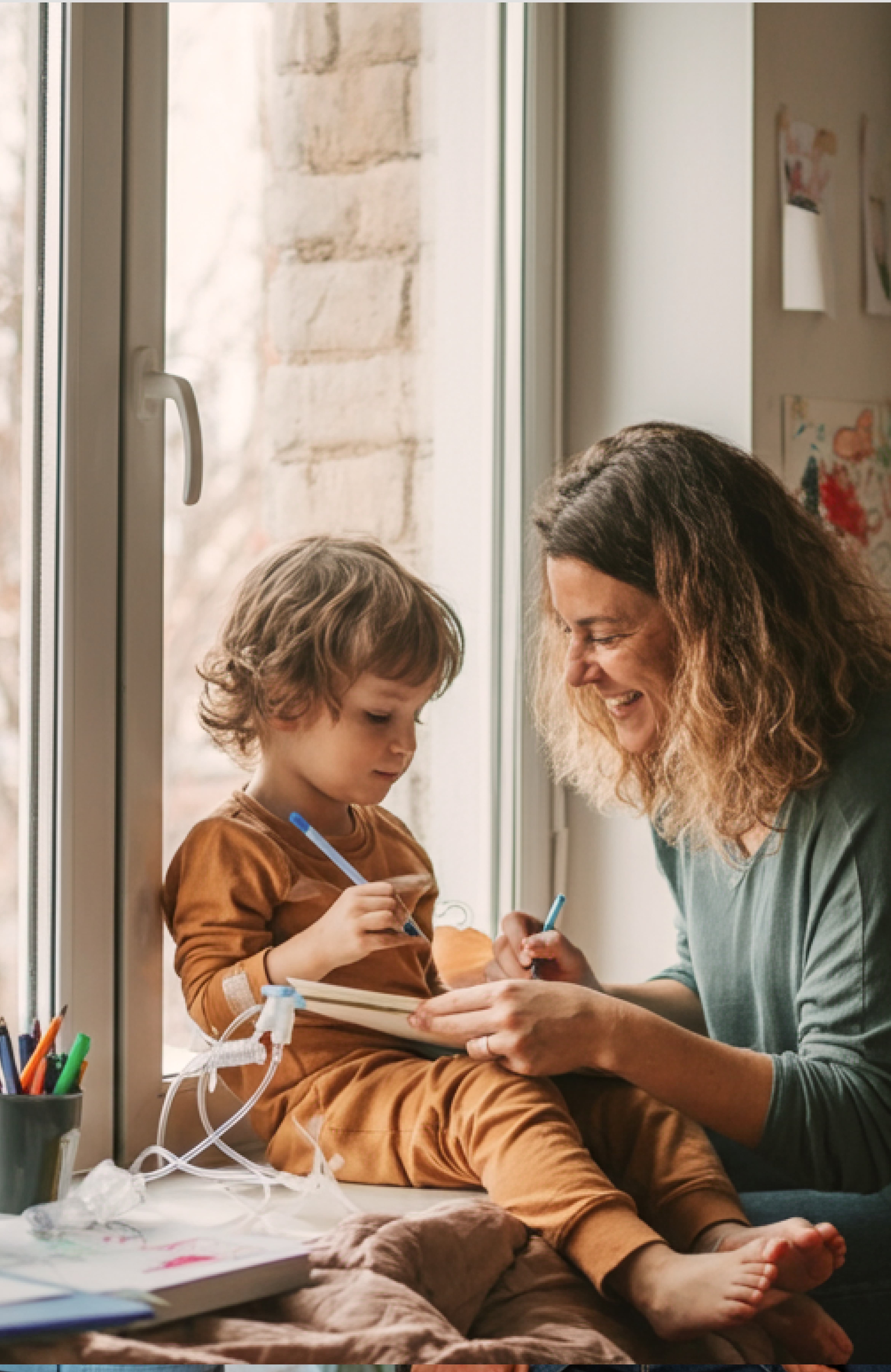 Child sitting by a window with an IV attached, drawing in a notebook with a woman smiling beside them.