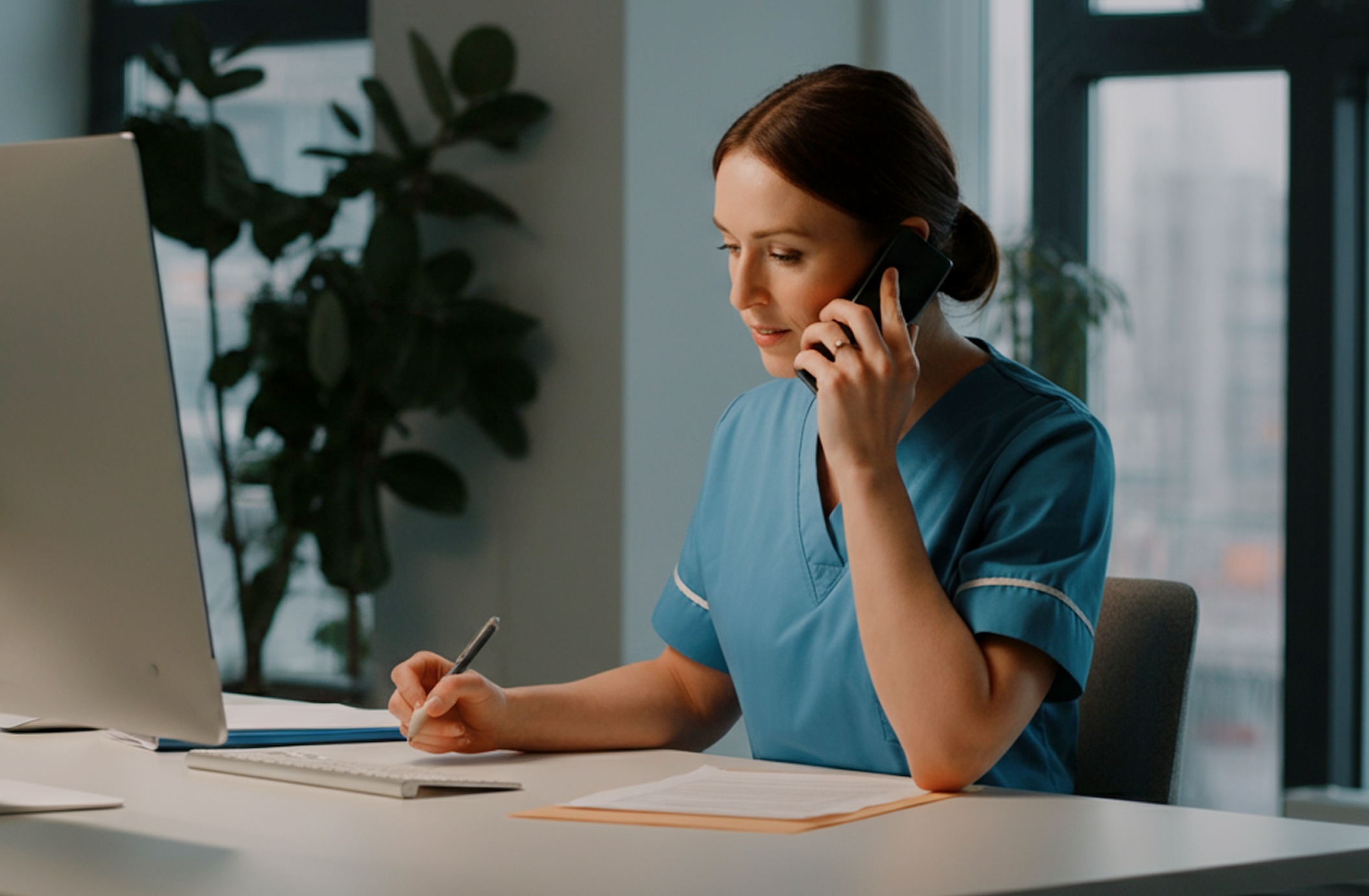 Nurse in blue scrubs talking on a phone while writing notes at a desk with a computer and documents.