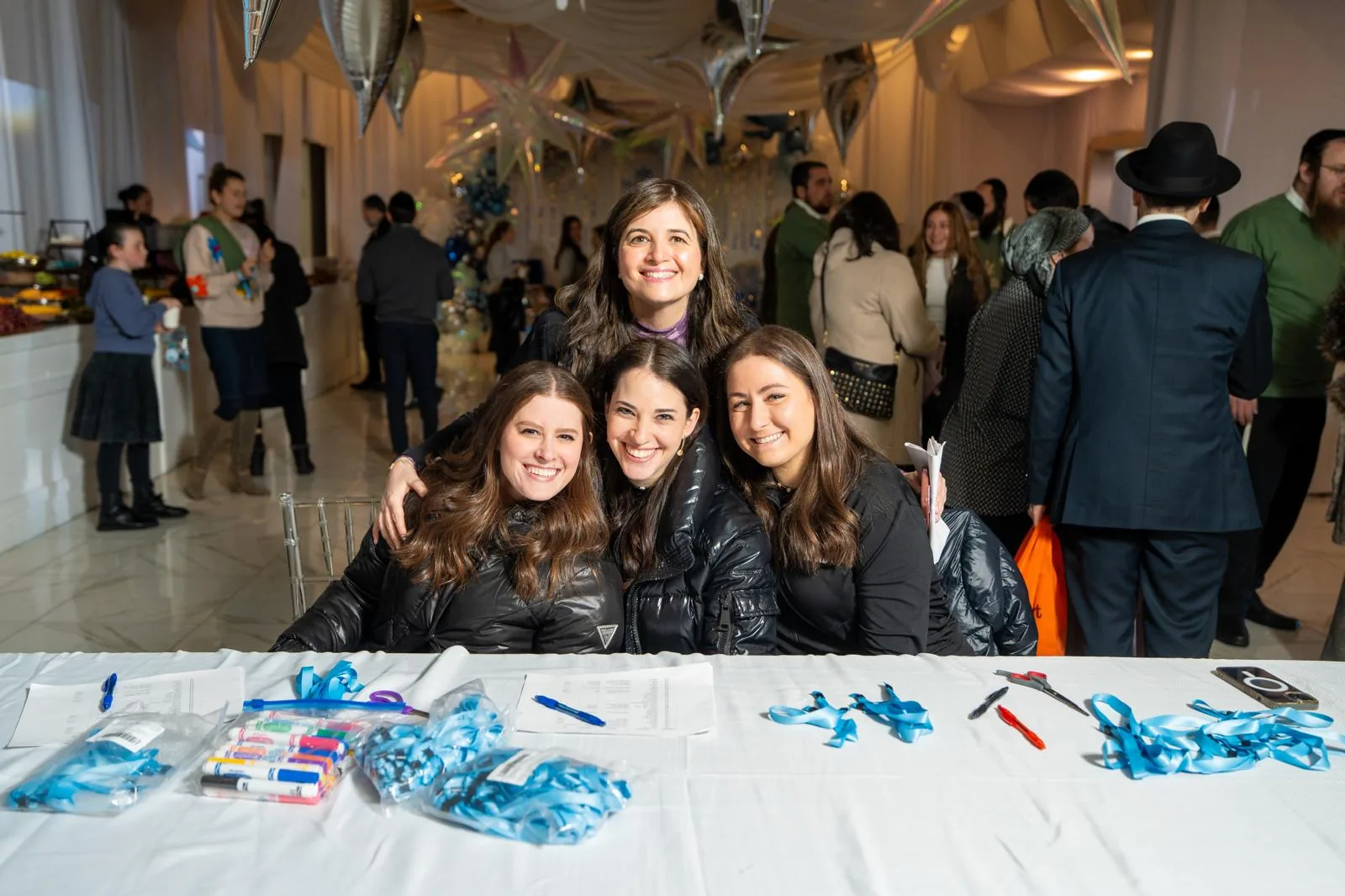 Four women smiling behind a table with ribbons and craft supplies at an event.