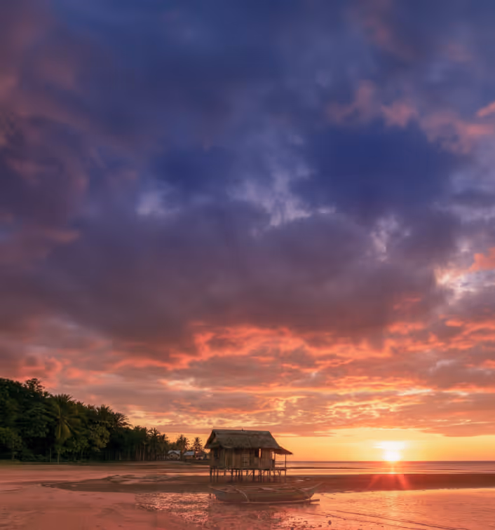 Sunset over beach with a small wooden hut on stilts and boat on sand near water.