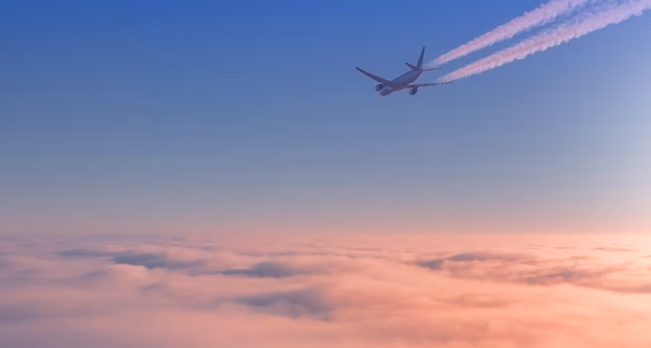 Airplane flying above pink clouds at sunset leaving contrails in a clear blue sky.