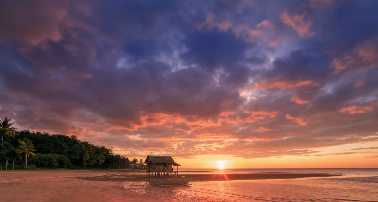 Sun setting over beach with palm trees, clouds, and a small hut on stilts near the water.
