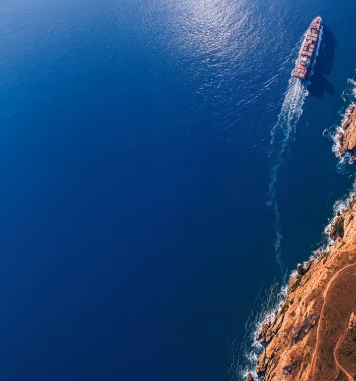 A cargo ship sailing near a rocky coastline with blue ocean water surrounding it.