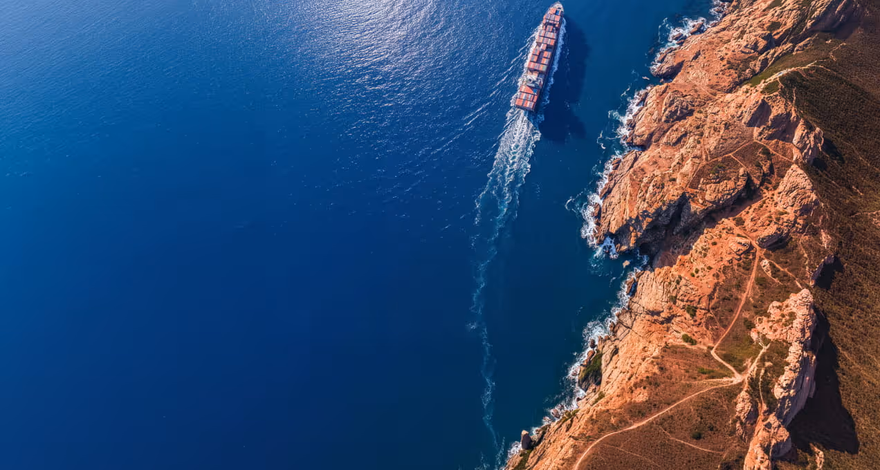 A cargo ship sailing along a rocky coastline with clear blue ocean water.