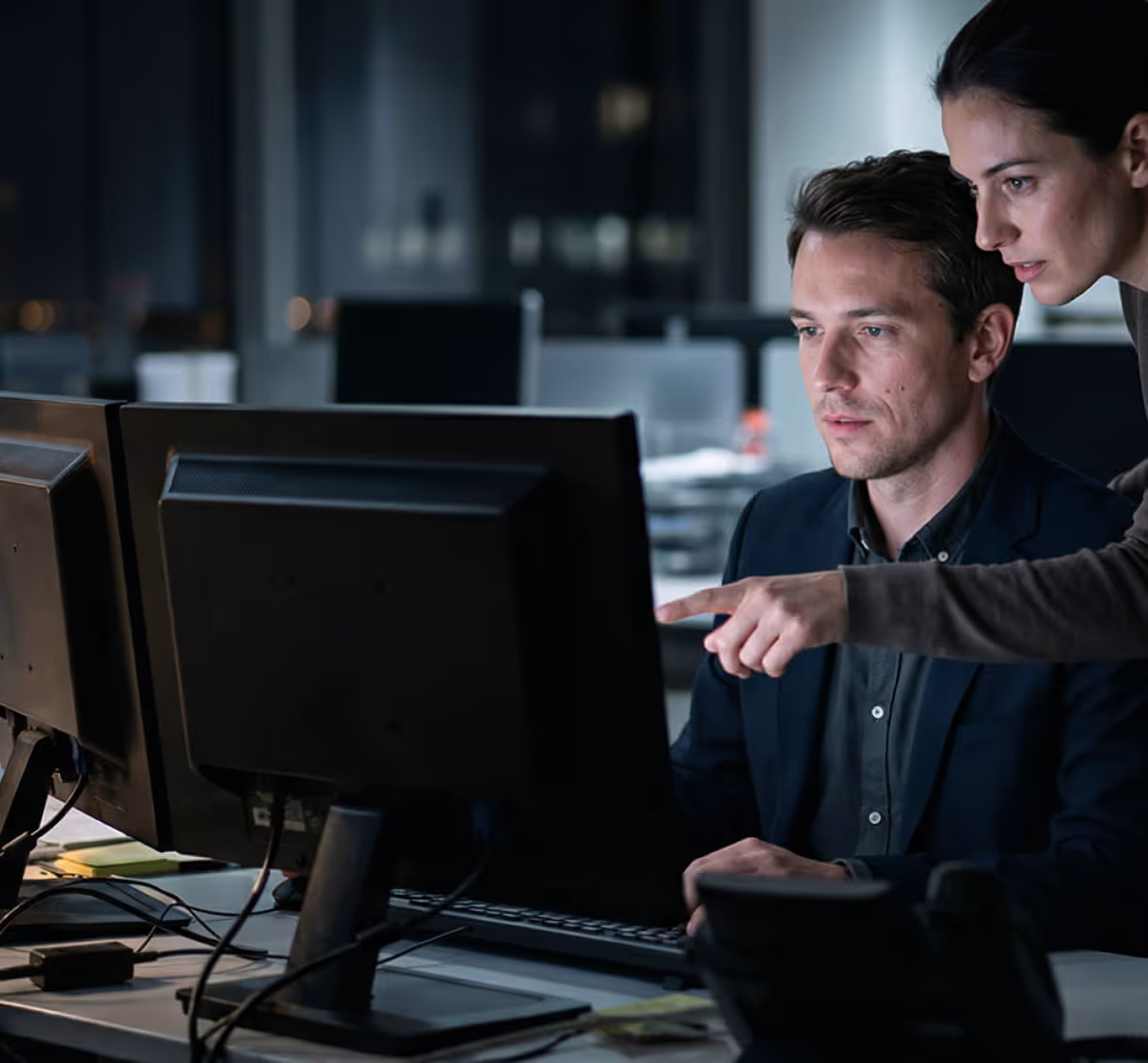 Woman points at computer screen while man attentively looks at it in a dark office.