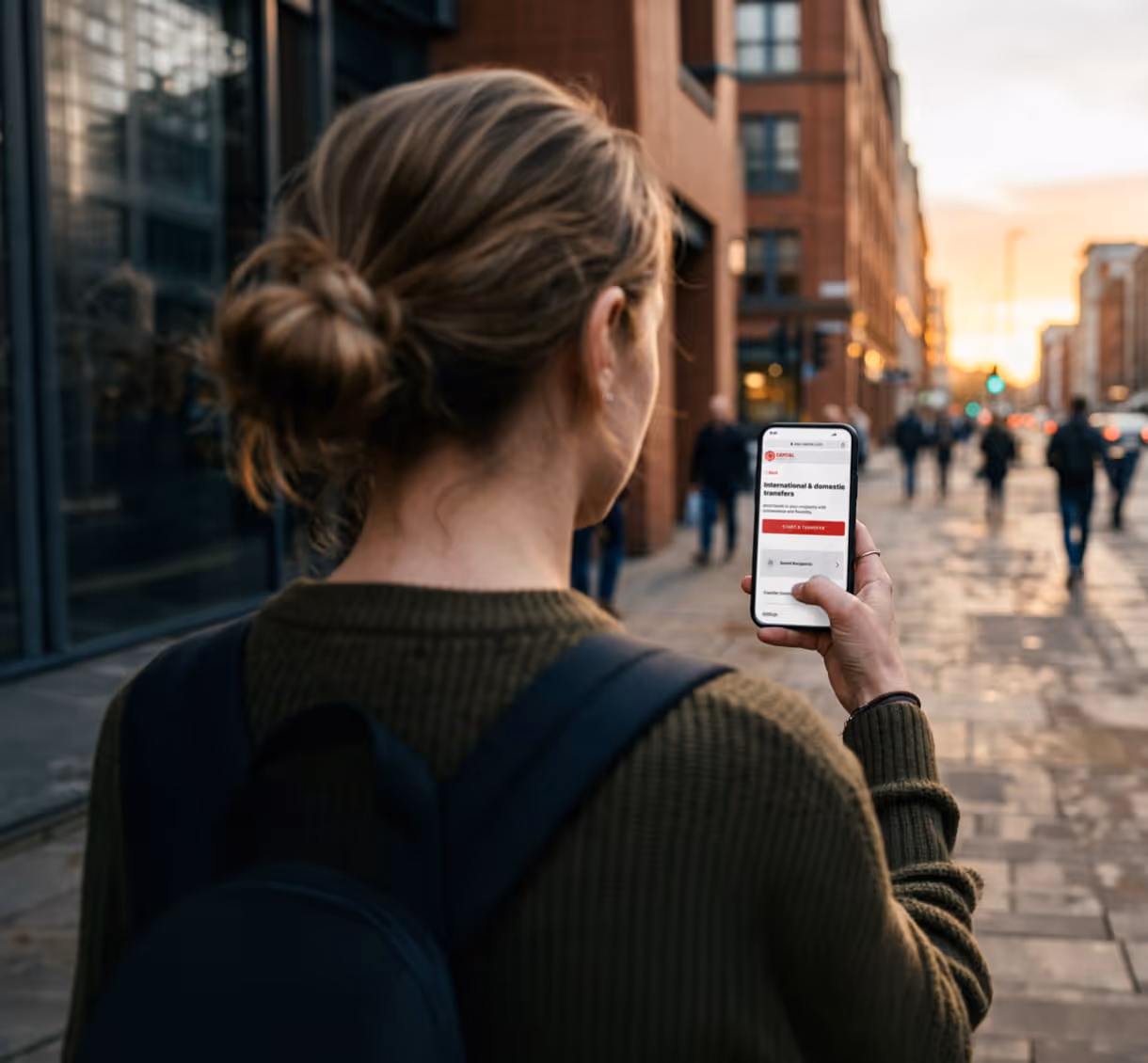 Woman using a smartphone on a city street at sunset, viewing an international and domestic transfers page.
