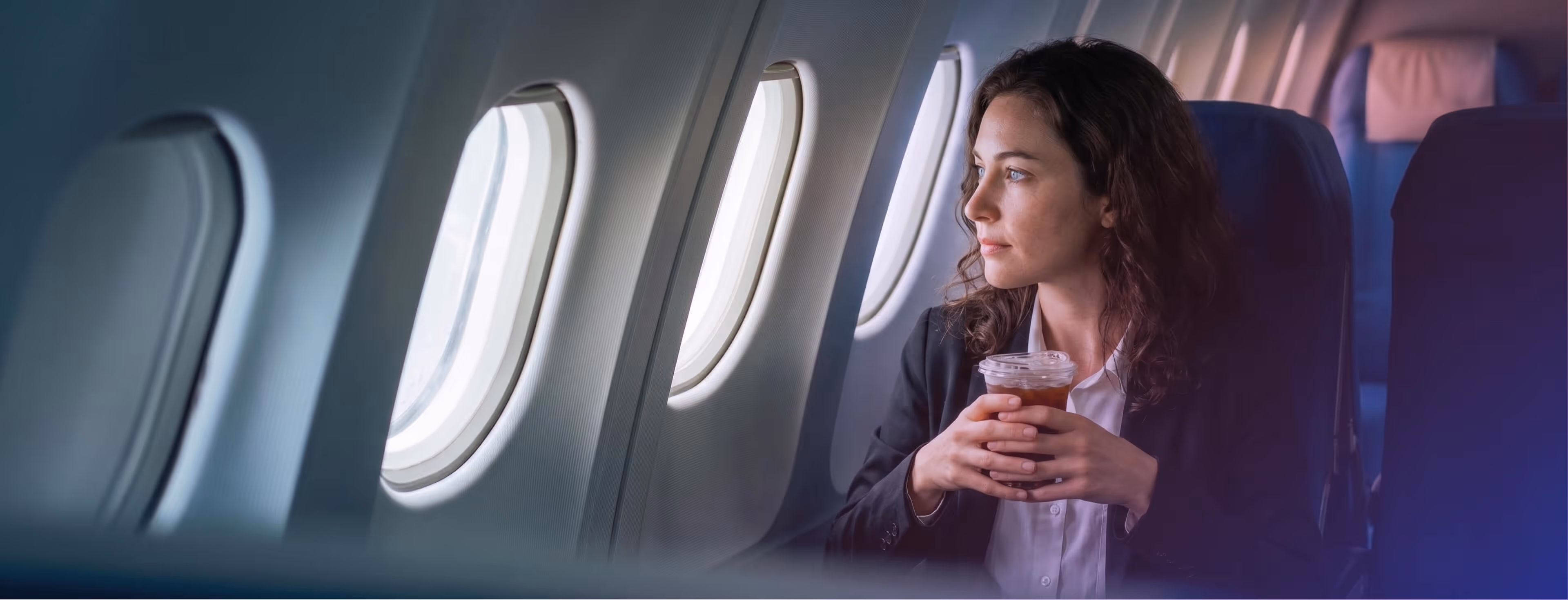 Woman holding a cold drink and looking out airplane window while seated in cabin.