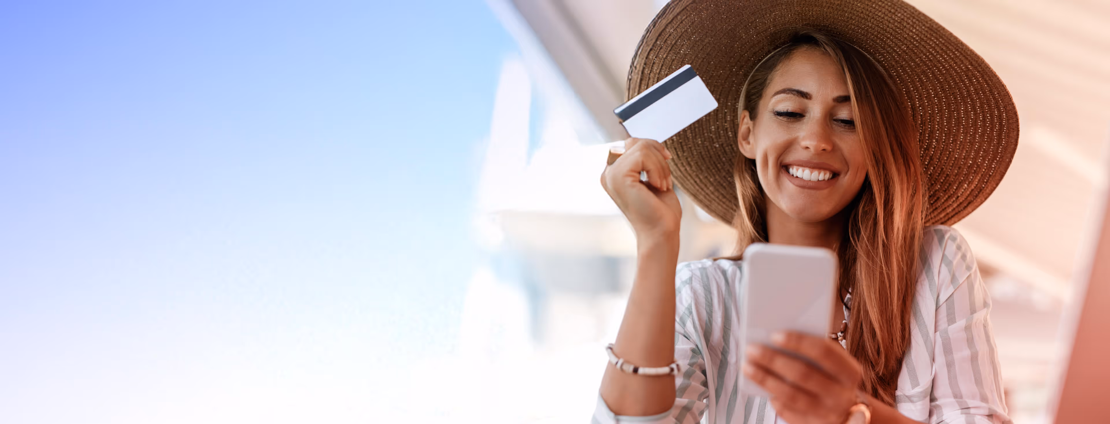 Smiling woman wearing a hat, holding a credit card and smartphone, likely doing online shopping.