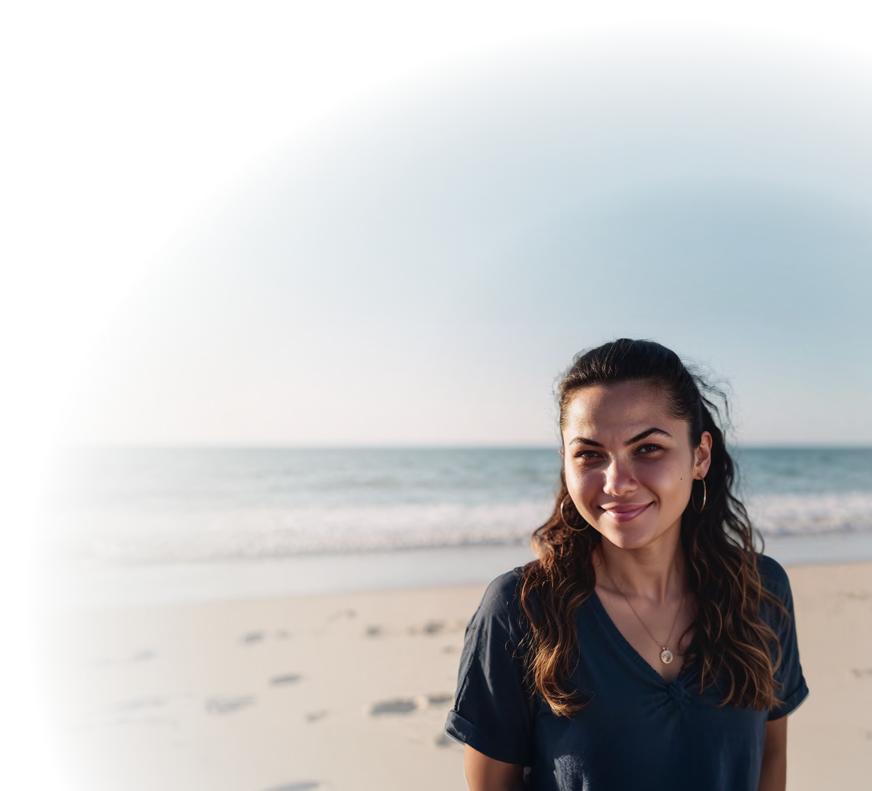 Young woman with long hair smiling on a sandy beach with ocean waves in the background.