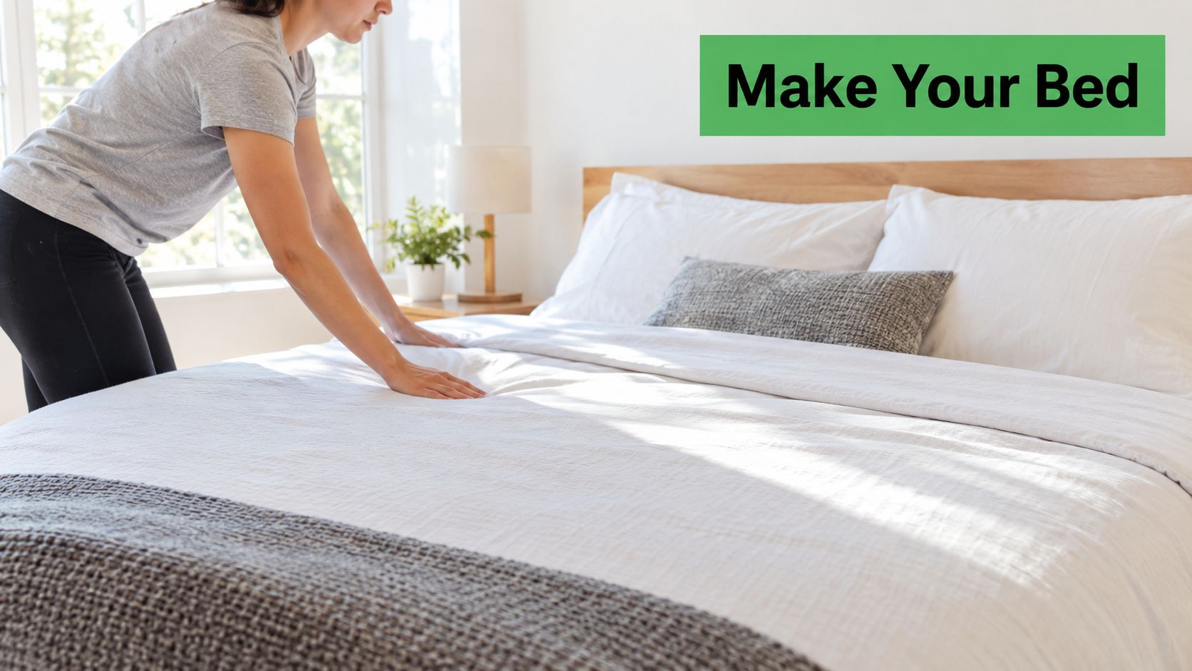 A woman making her bed by smoothing the white duvet in a bright, modern bedroom.
