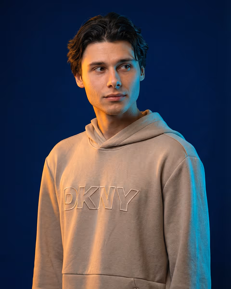 Чоловік у бежевій толстовці з капюшоном і написом DKNY на грудях дивиться вправо на темно-синьому фоні.