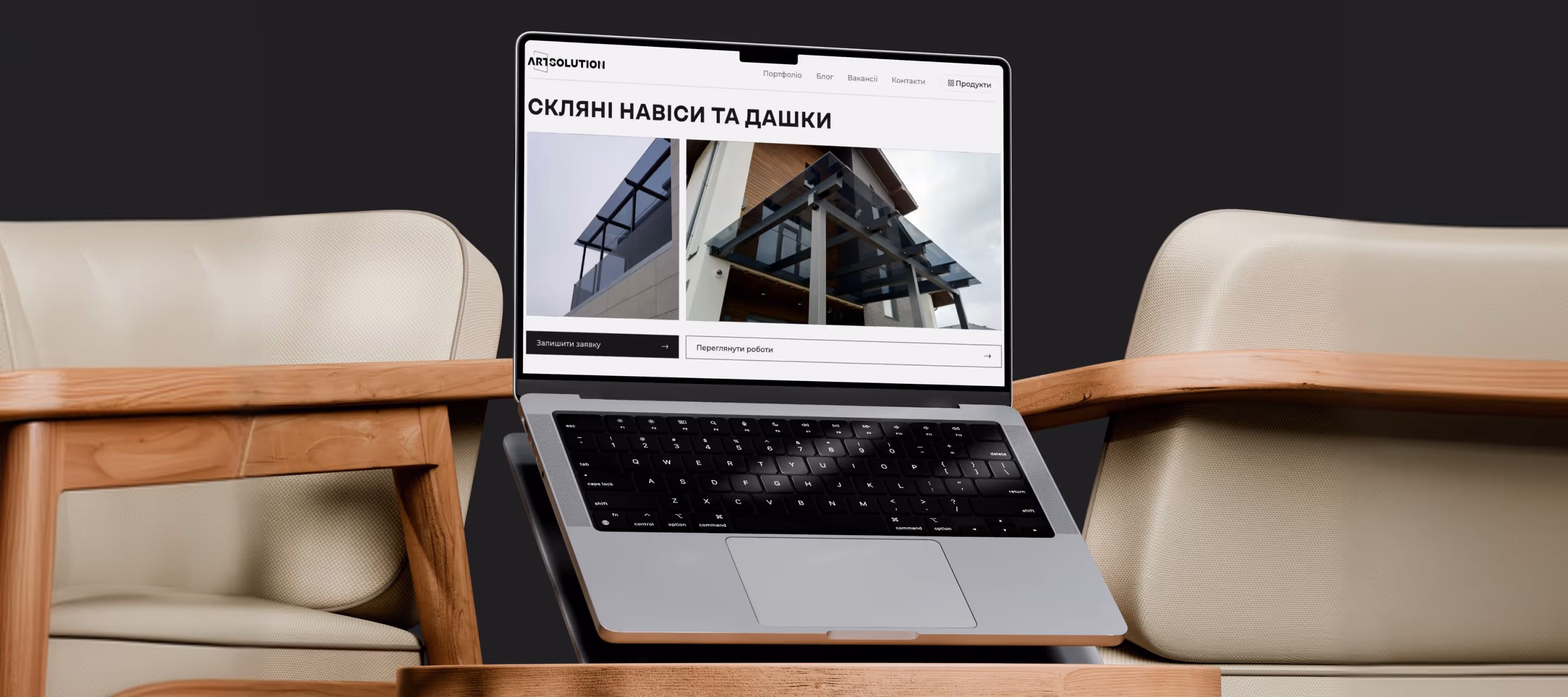 Ноутбук із відкритою веб-сторінкою компанії Art Solution, що показує скляні навіси та дашки, розміщений між двома бежевими кріслами з дерев'яними підлокітниками.