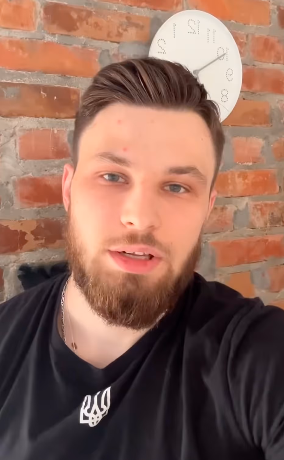 Чоловік із сивим волоссям і бородою на фоні цегляної стіни і круглого годинника.