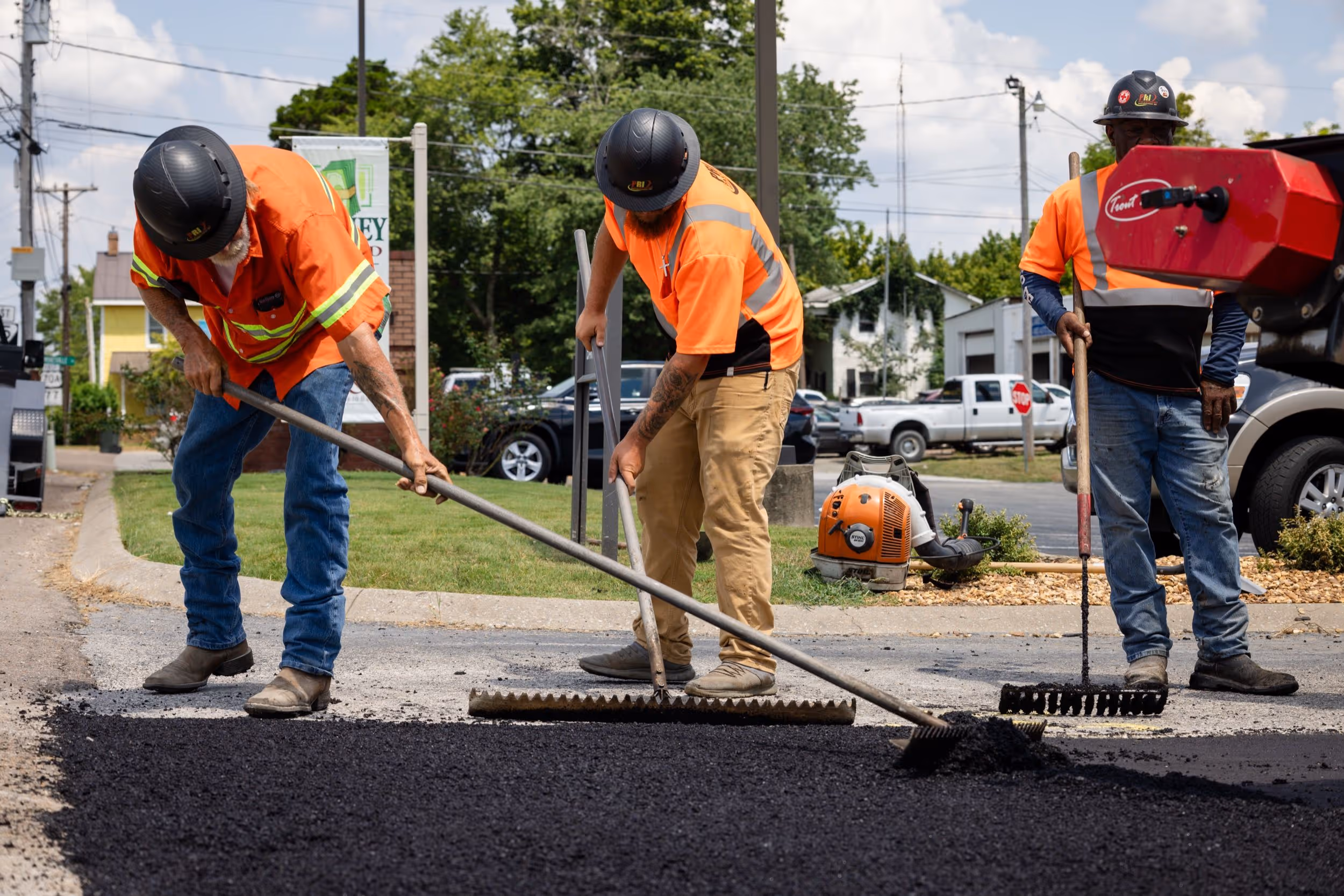 Three construction workers in orange safety shirts spreading fresh asphalt on a road.