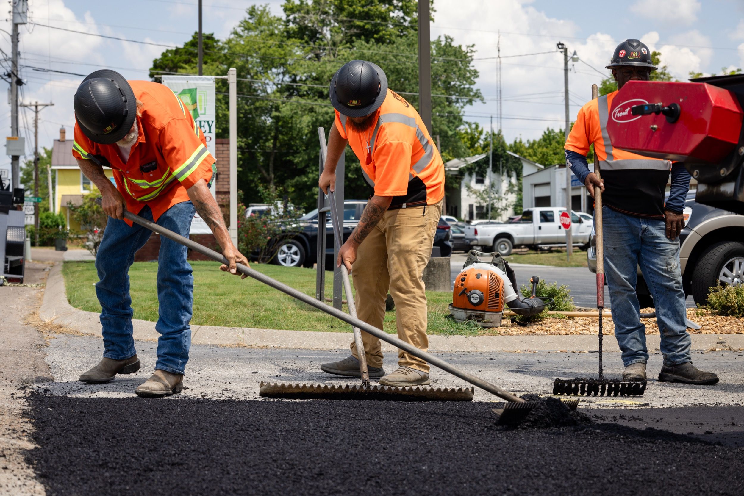 Three construction workers in orange safety shirts spreading fresh asphalt on a road.