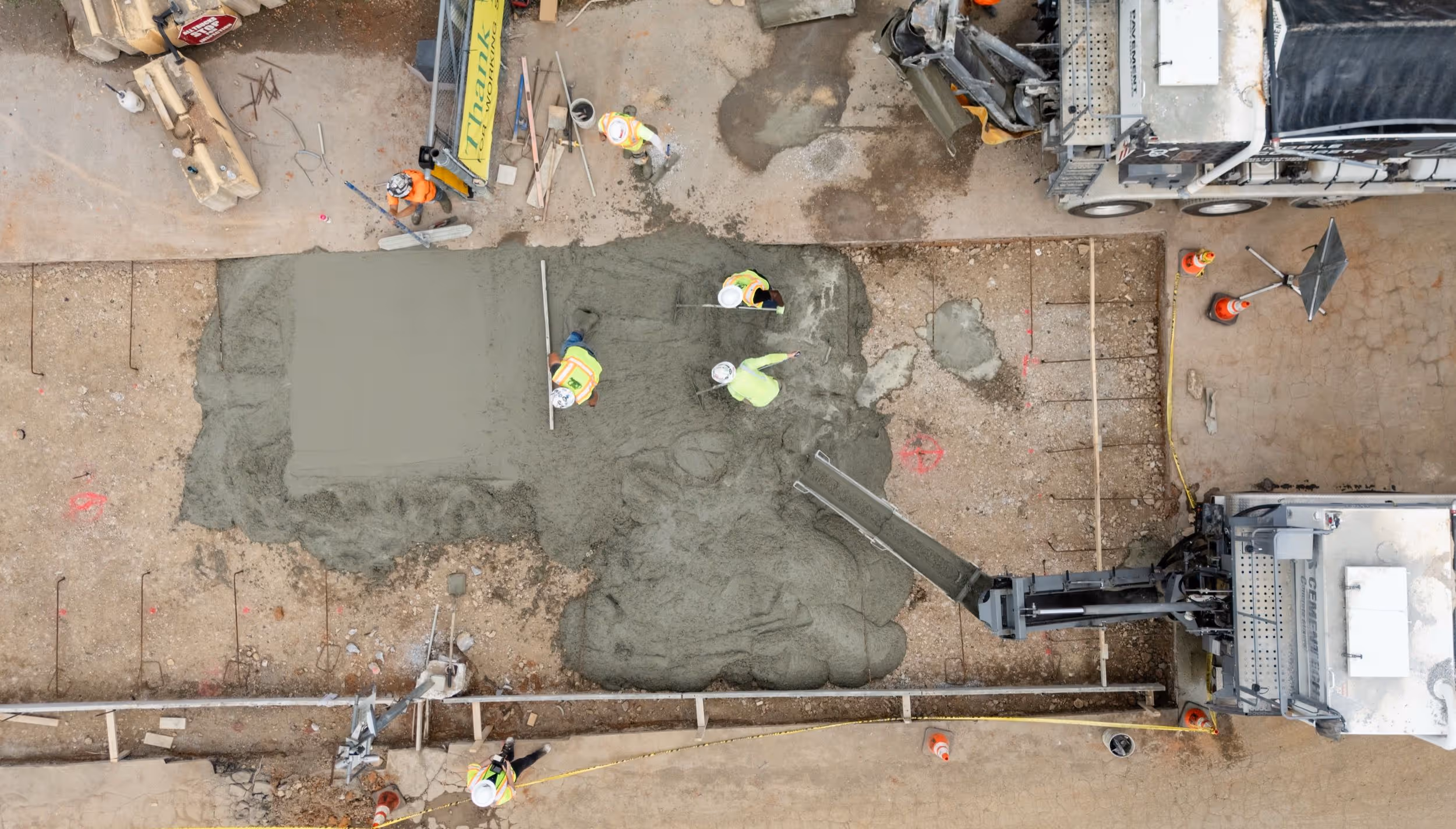 Aerial view of construction workers spreading and smoothing wet concrete on a ground surface near a concrete mixer truck.