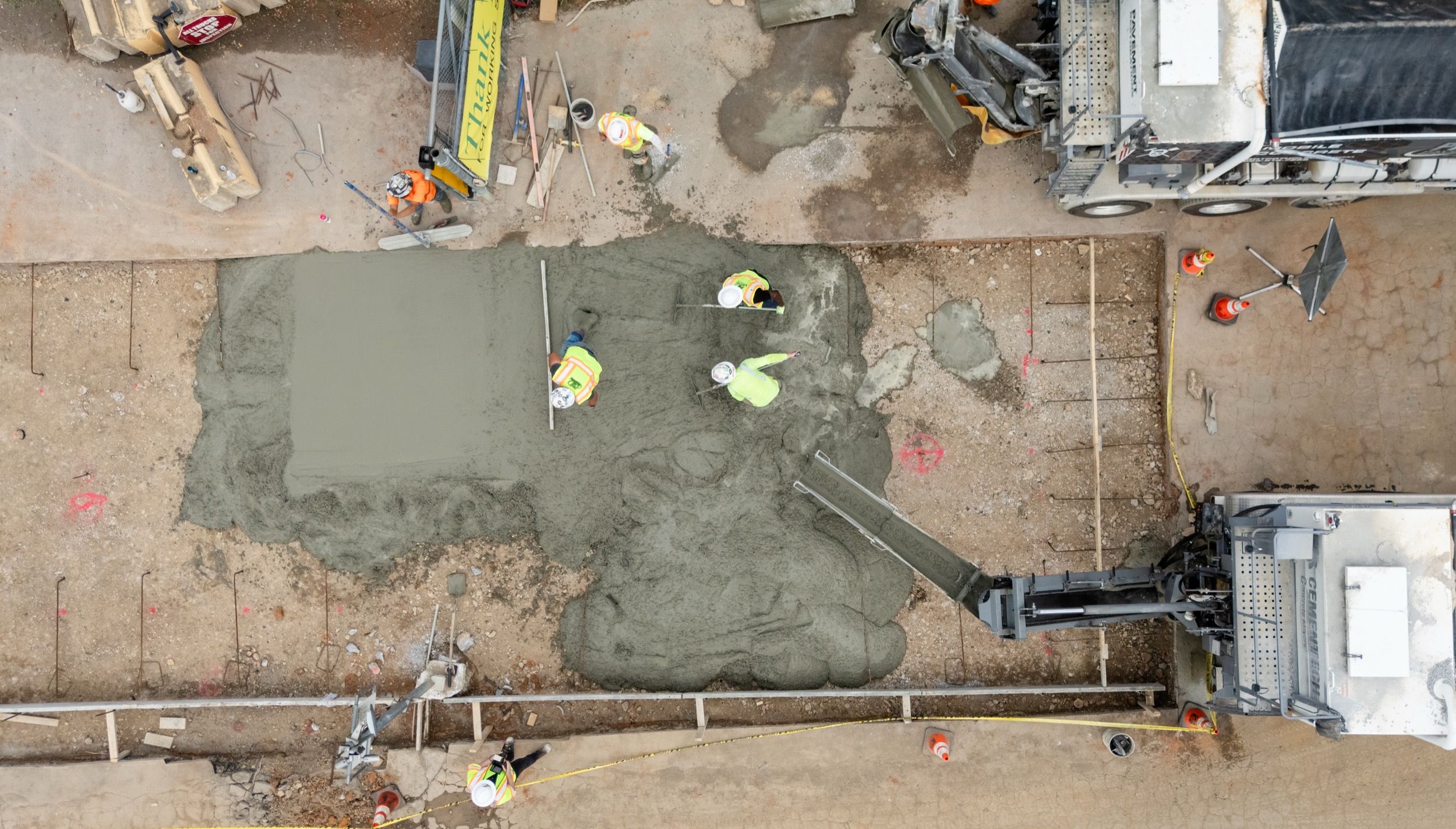 Aerial view of construction workers spreading and smoothing wet concrete on a ground surface near a concrete mixer truck.
