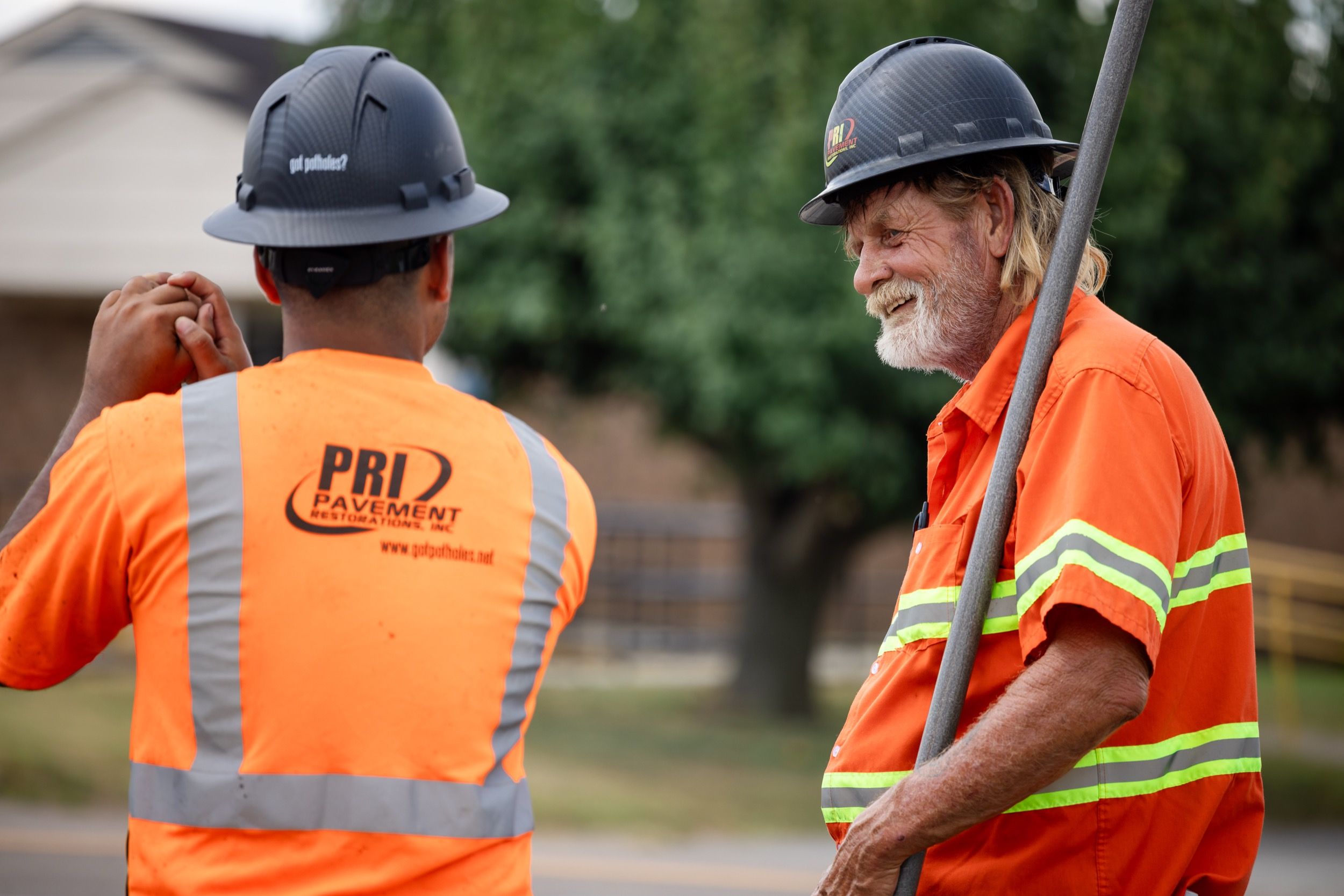 Two construction workers in orange safety shirts and black helmets talking outdoors near a tree.