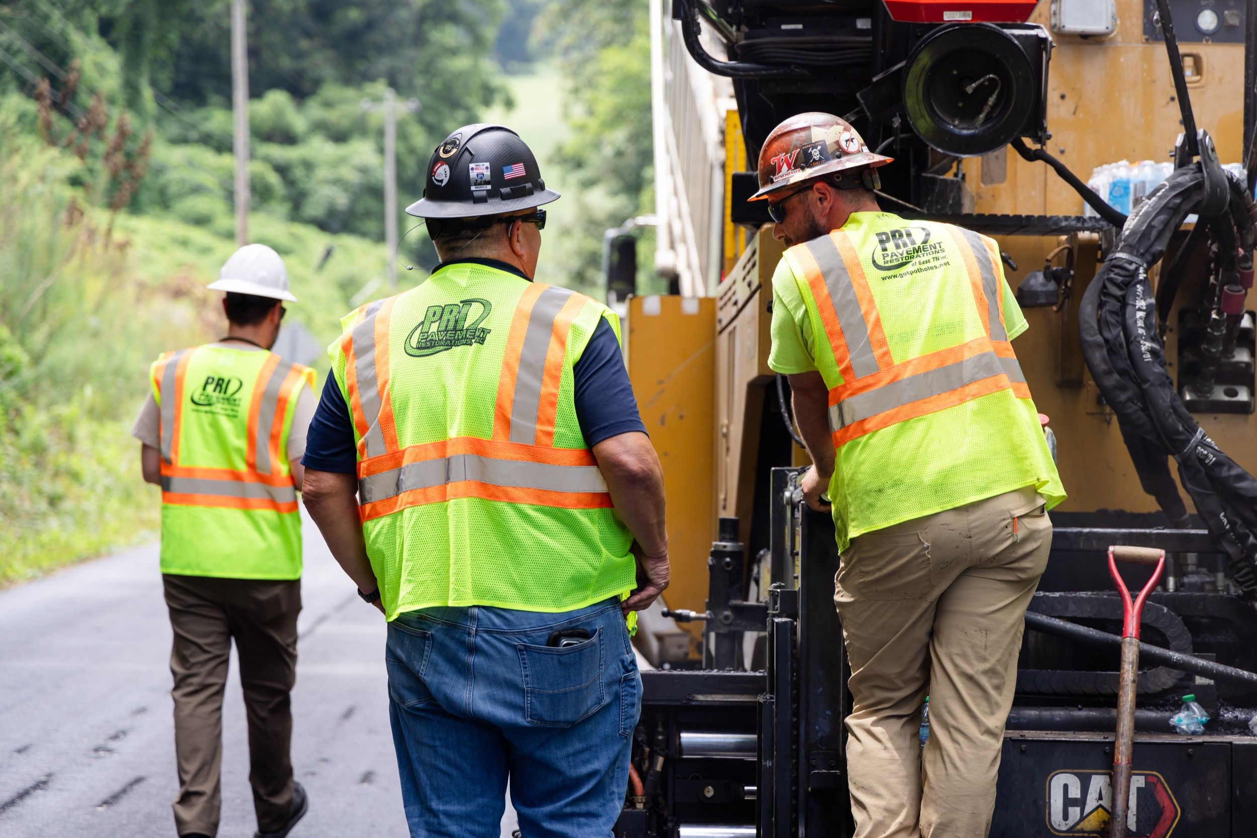 Three road workers wearing high-visibility vests and hard hats operating paving machinery on a rural road.