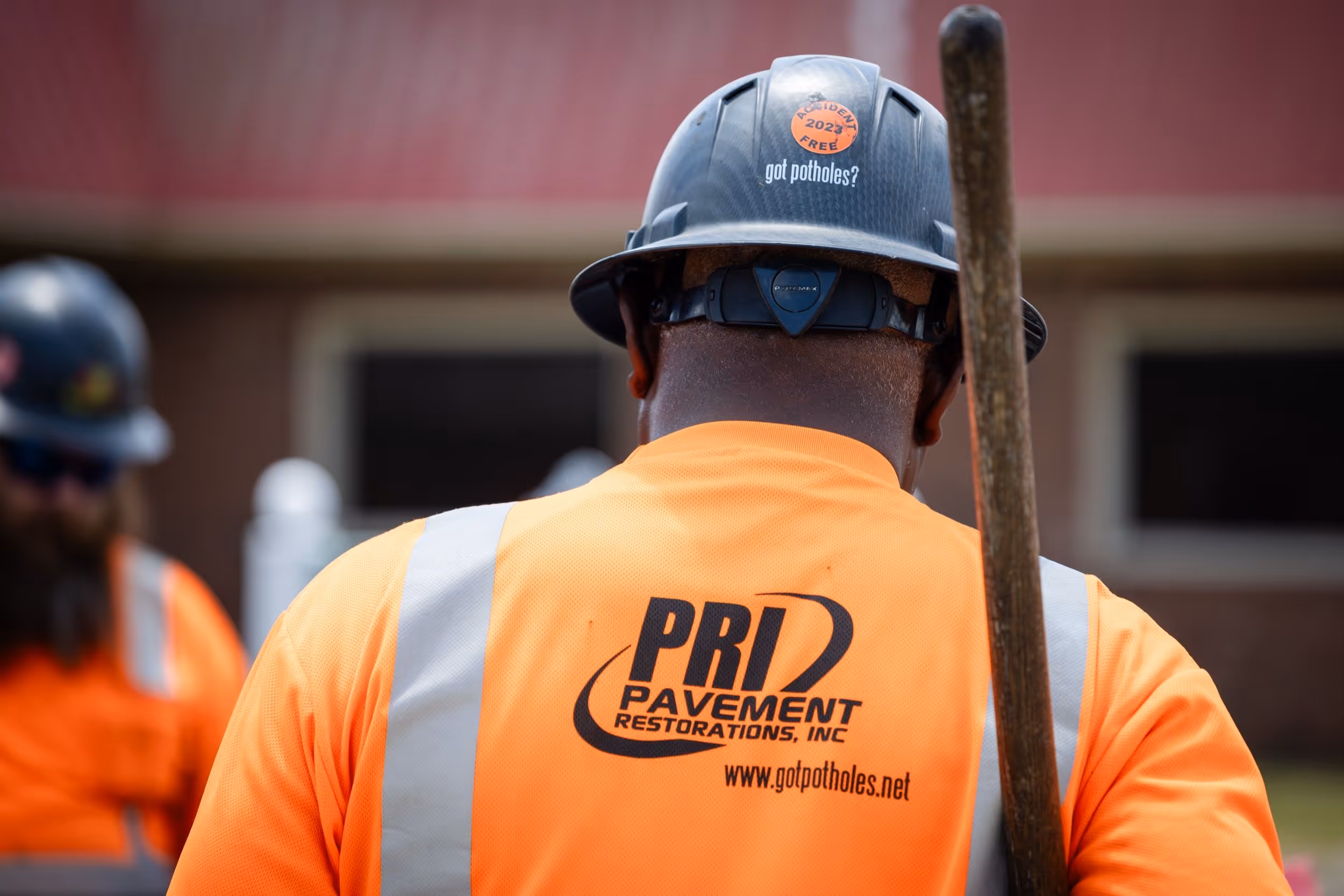 Worker wearing an orange safety shirt and black hard hat with 'got potholes?' text and PRI Pavement Restorations logo on shirt back.