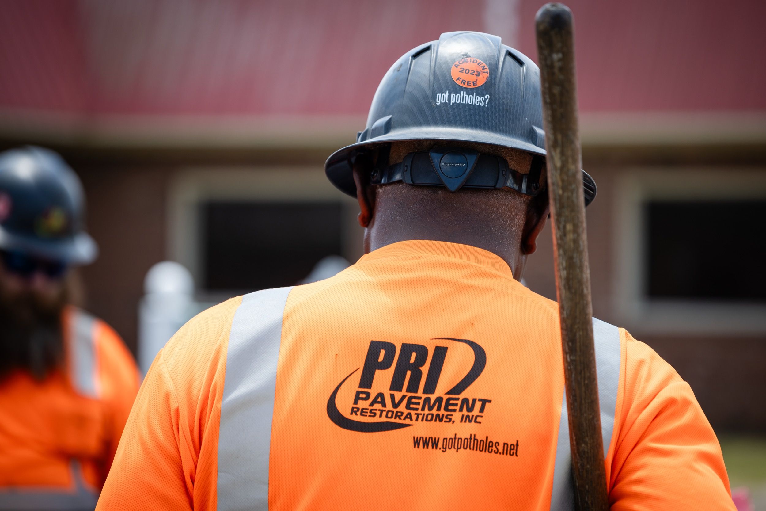 Worker wearing an orange safety shirt and black hard hat with 'got potholes?' text and PRI Pavement Restorations logo on shirt back.