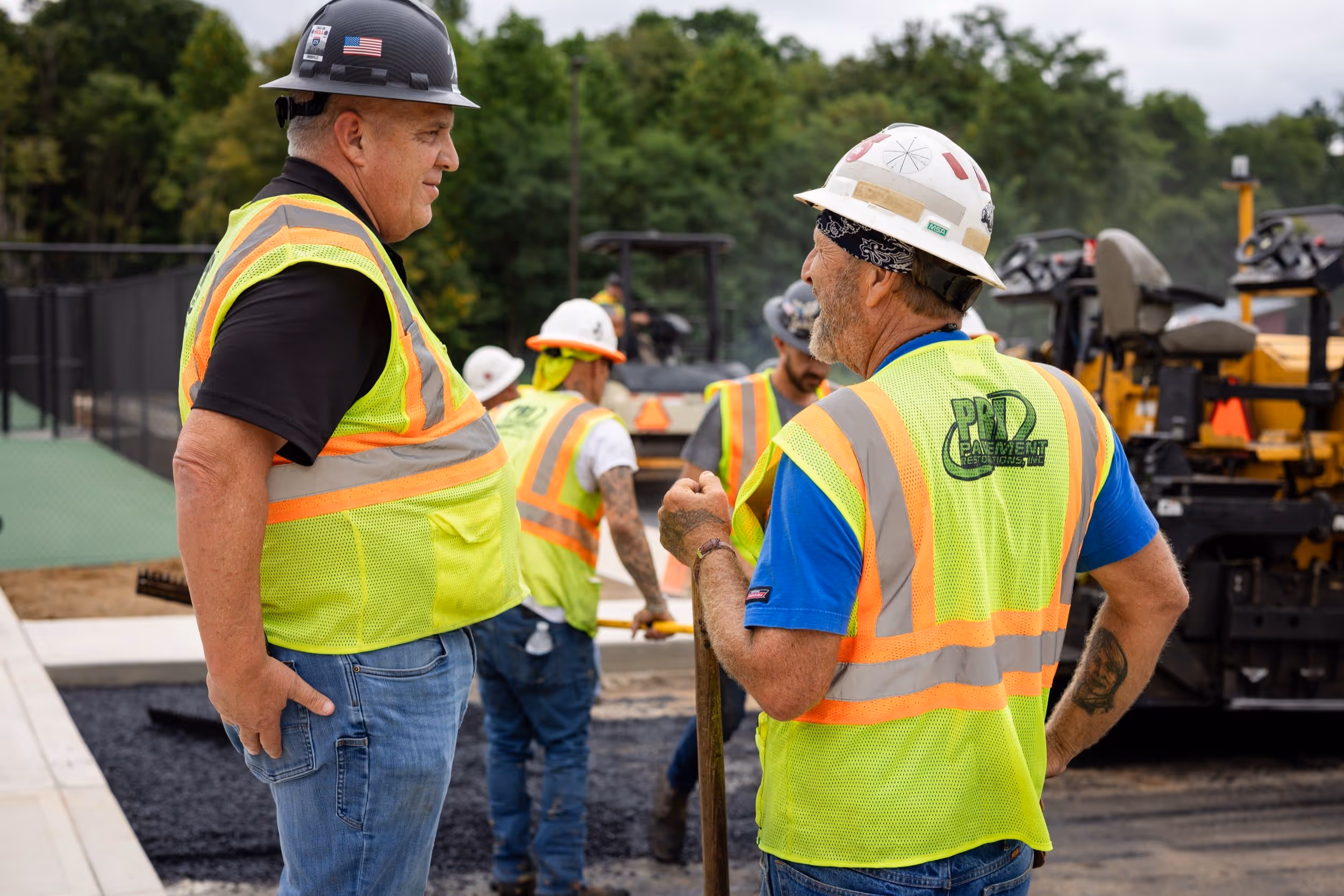 Construction workers in safety vests and helmets conversing at an asphalt paving site with equipment in the background.