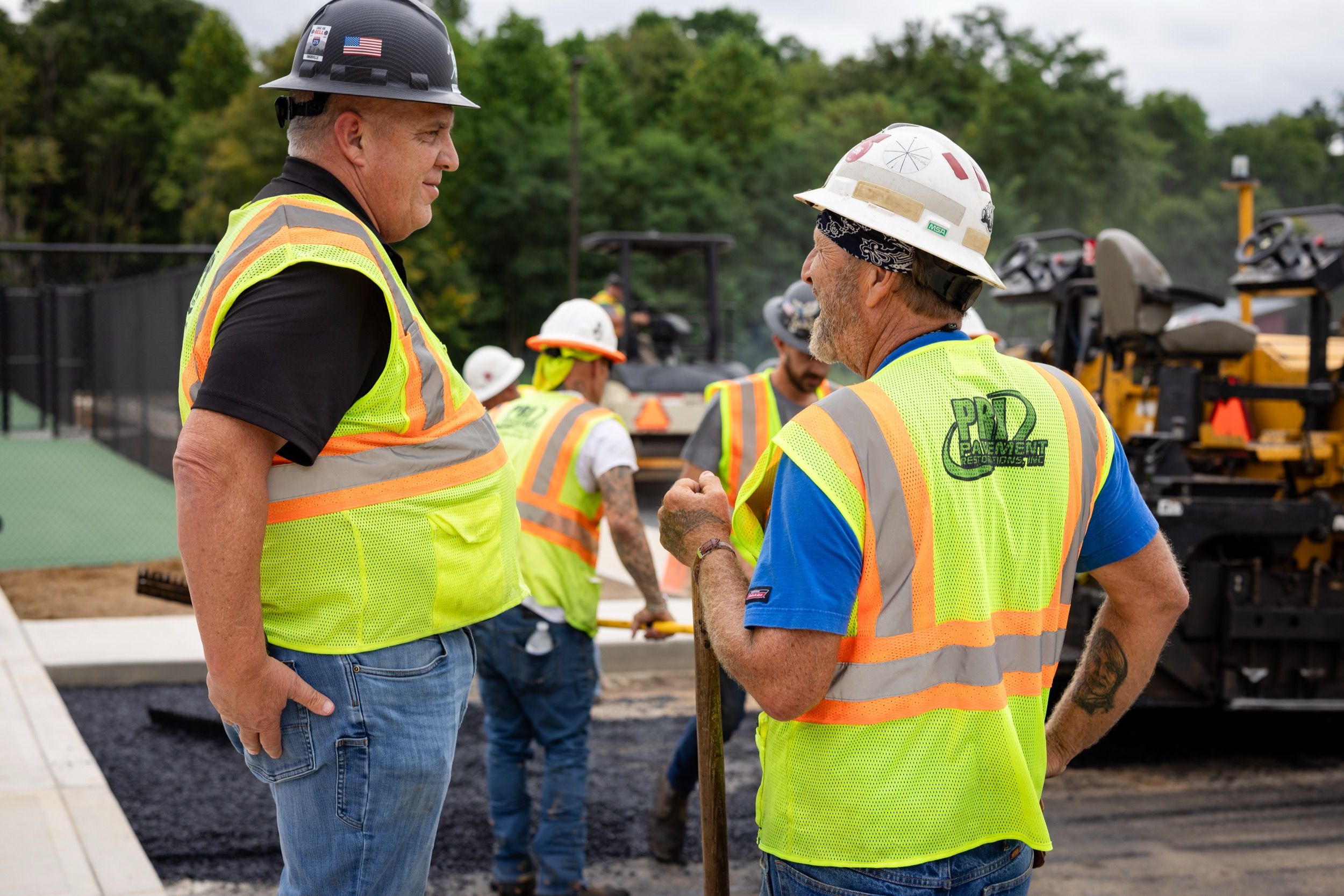 Construction workers in safety vests and helmets conversing at an asphalt paving site with equipment in the background.