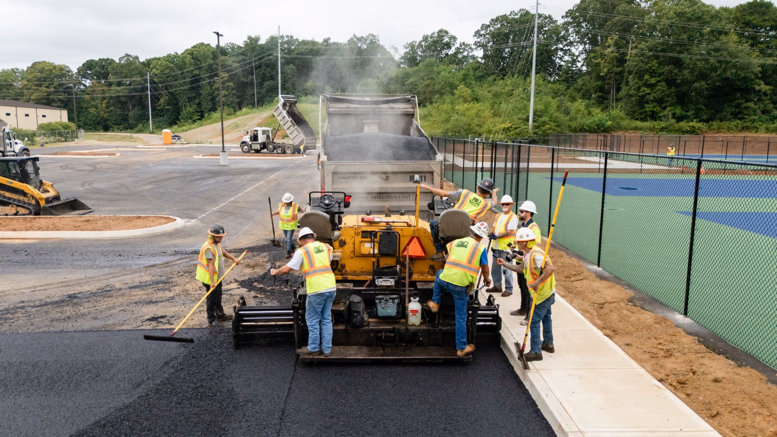 Construction workers in safety vests and helmets spreading fresh asphalt with machinery and tools on a parking lot near a fenced sports court.