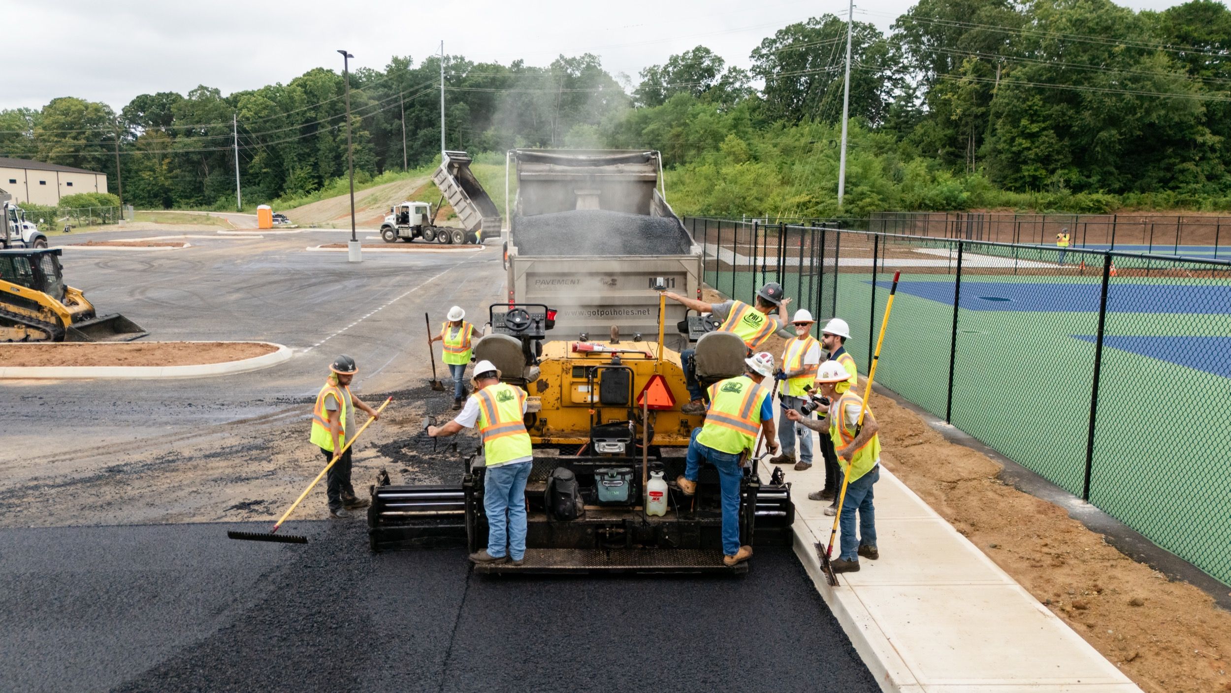 Construction workers in safety vests and helmets spreading fresh asphalt with machinery and tools on a parking lot near a fenced sports court.